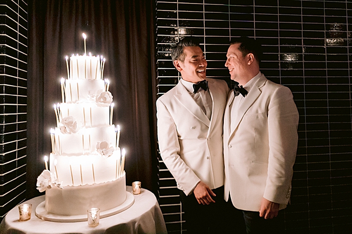 grooms smile at each other beside a wedding cake with lit candles