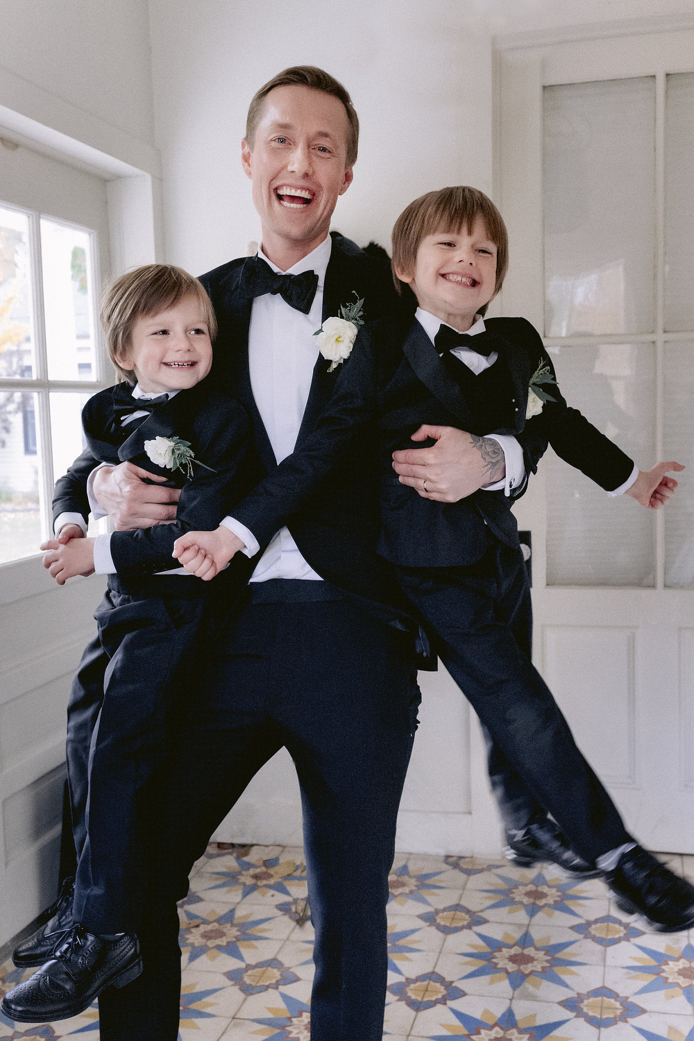 Happy groom horsing around with the two little ring bearers, smiling and laughing, all wearing matching tuxedos. Editorial Wedding at Firefox Mountain House in Upstate New York. 