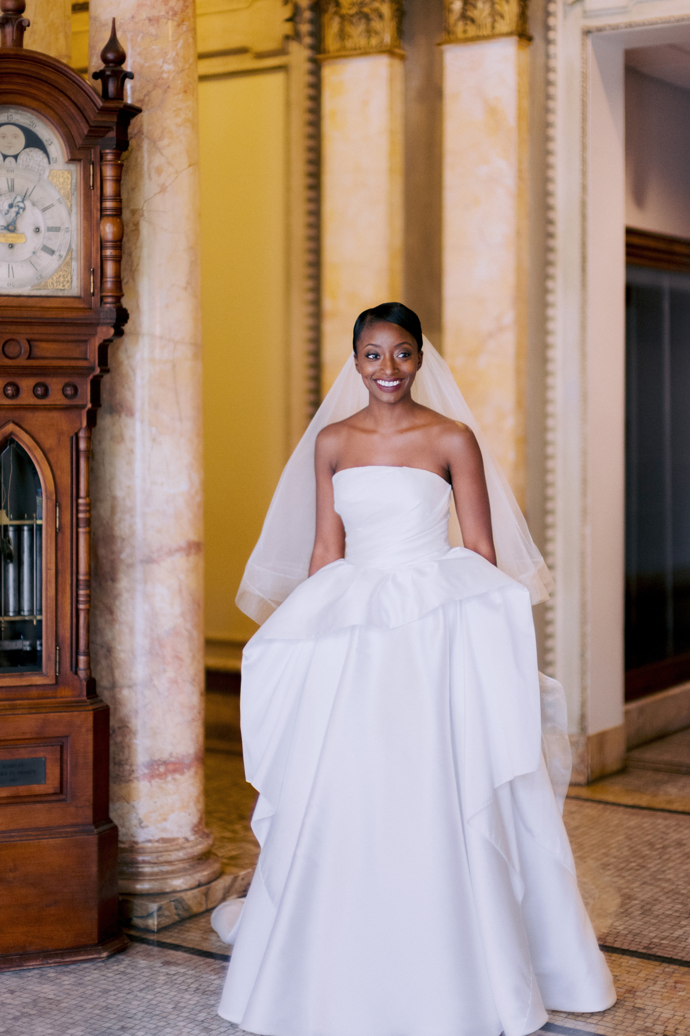 Bride wearing wedding dress standing by grandfather clock in Providence Public Library