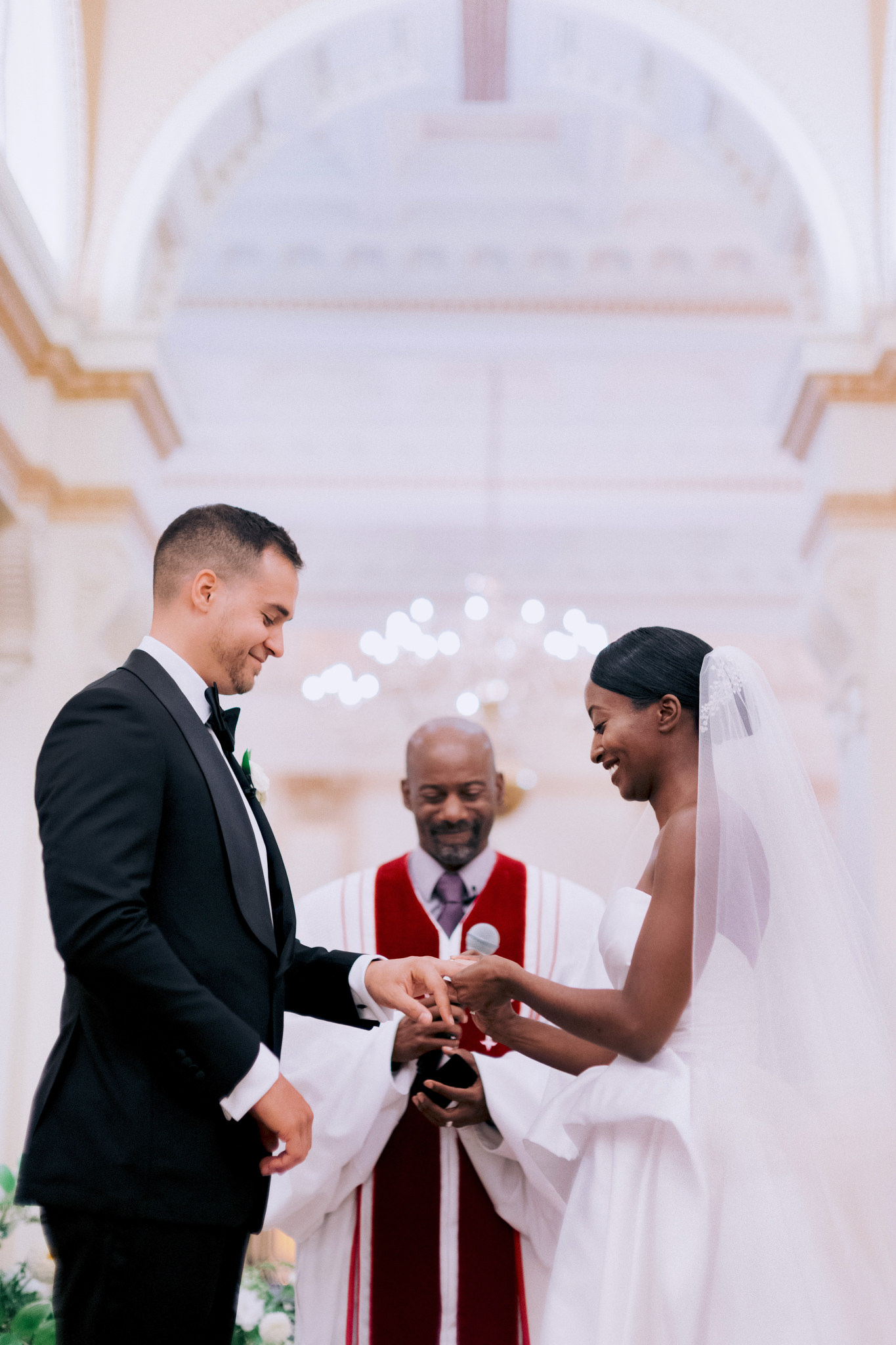 Interracial couple smiling on their wedding day as bride puts ring on grooms finger