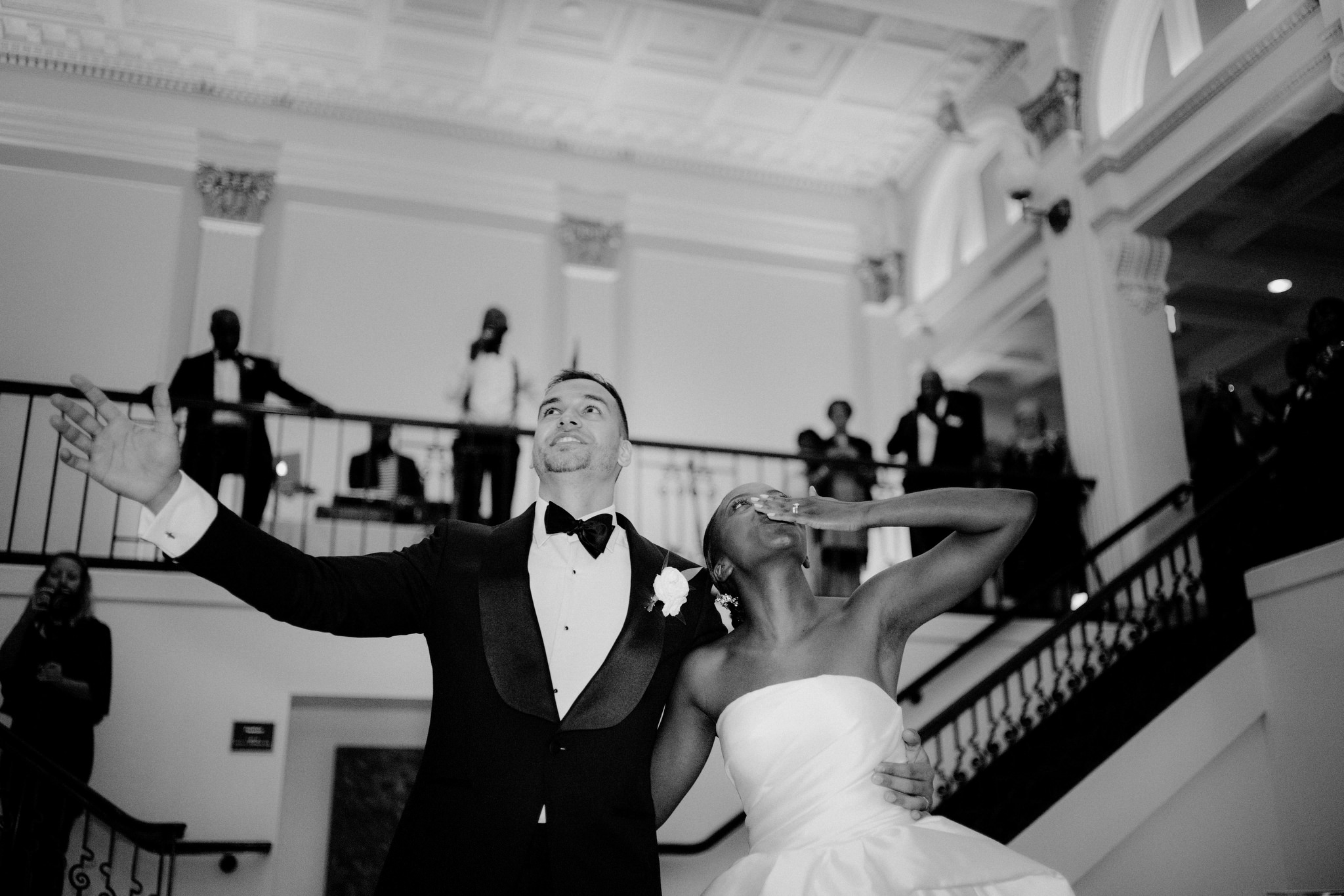 Couple with an arm around each other, waving to their guests as they descend the stairs for the first time as a married couple