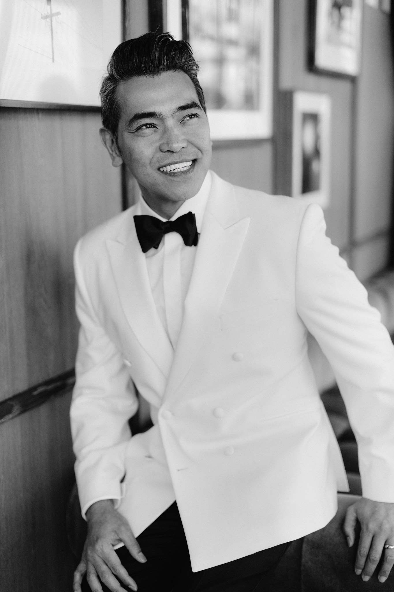 Man smiling wearing white suit with black bow tie and wedding band on his finger