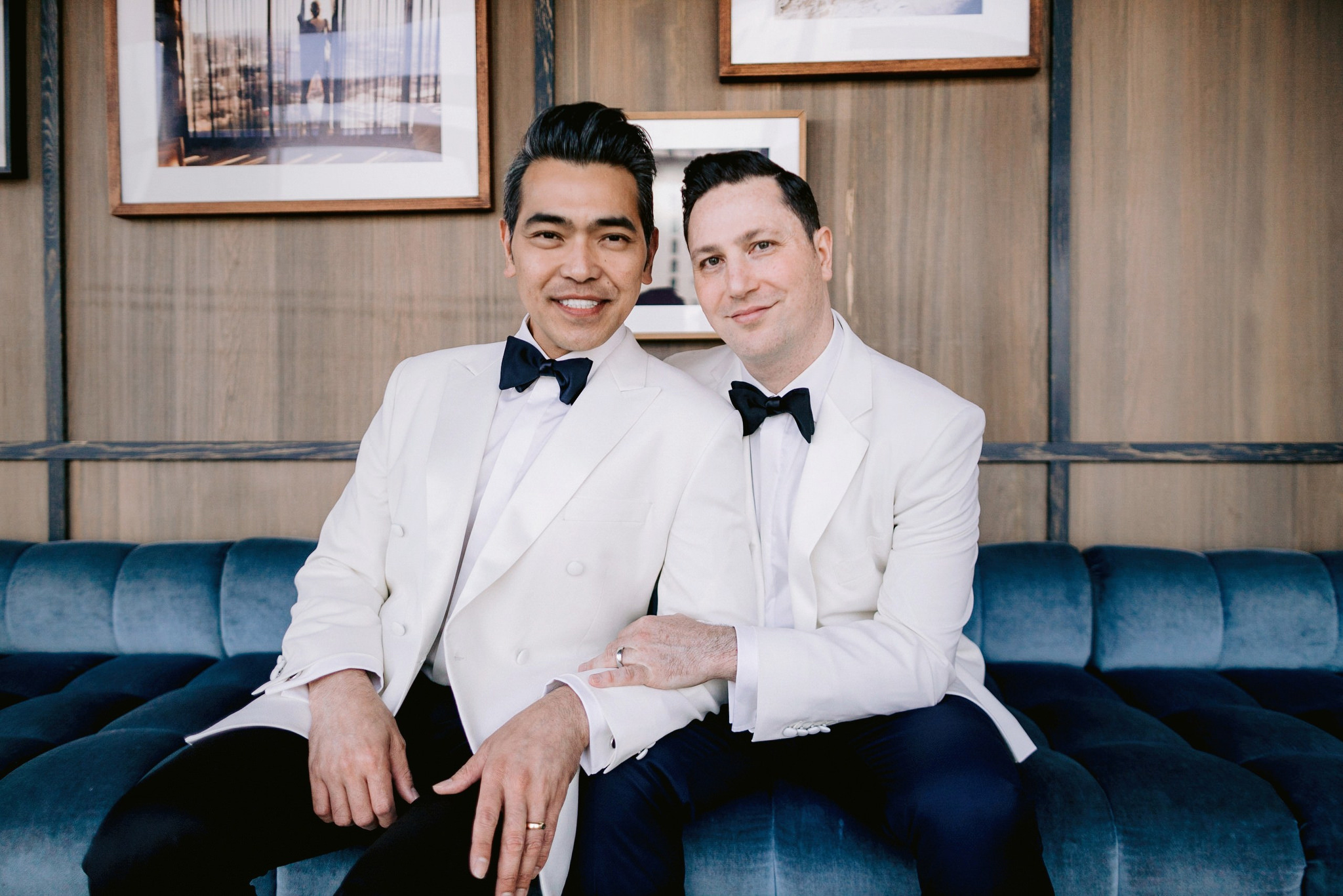 Same sex male couple sitting together smiling wearing white suits with black bow ties for their wedding
