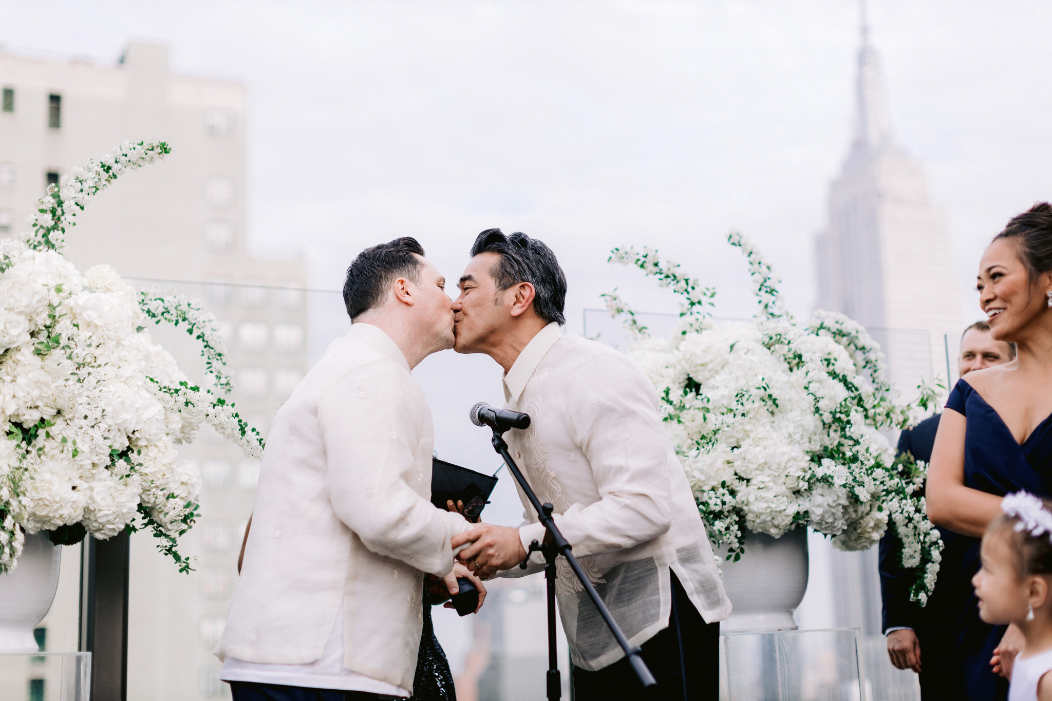 Same sex male couple kiss after saying I do in white suits at rooftop wedding
