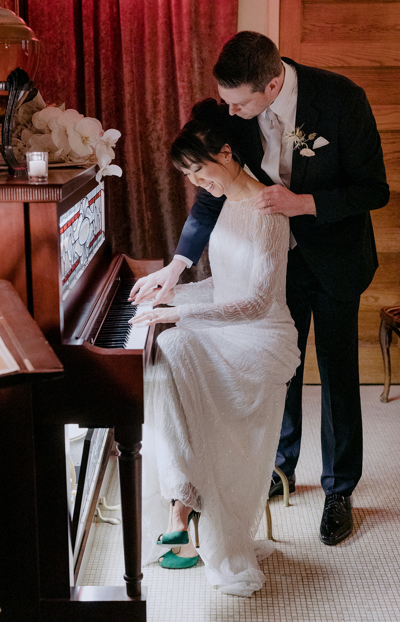 Men reaches over womans shoulder as she plays piano in wedding dress