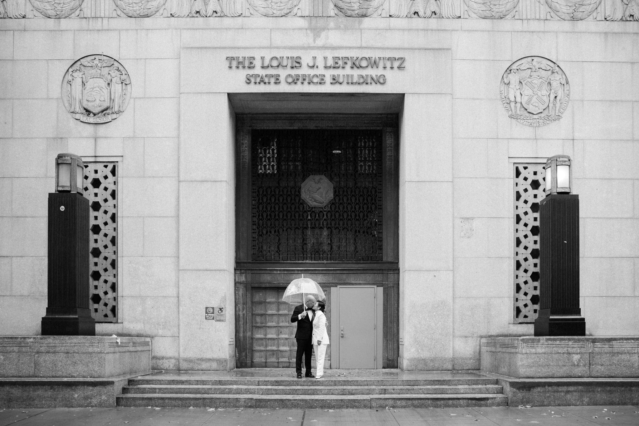 Bride and groom kiss under an umbrella outside of the Louis J Lefkowitz State Office Buildling.