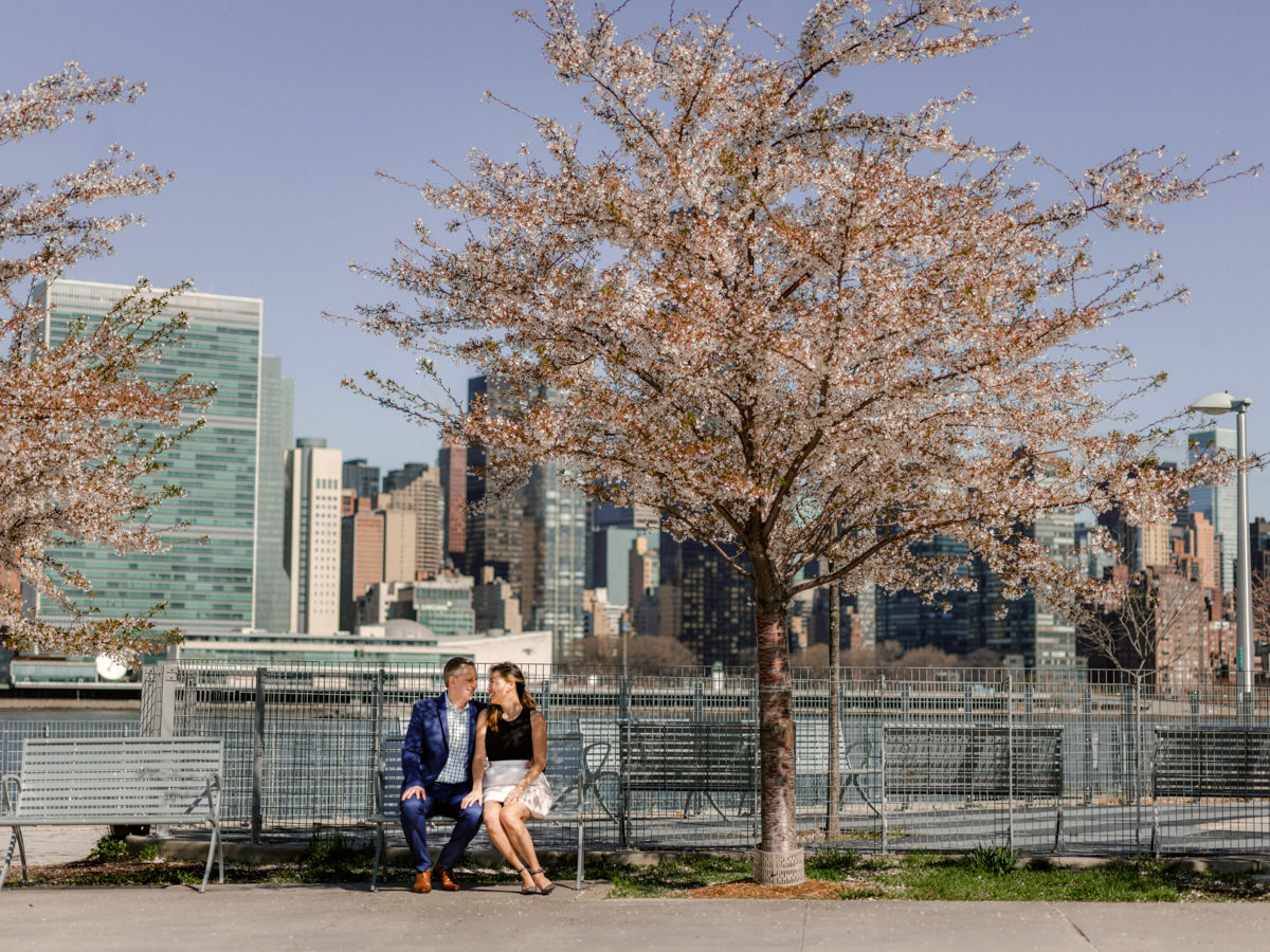 An engagement couple sits on a park bench with the New York City skyline behind them