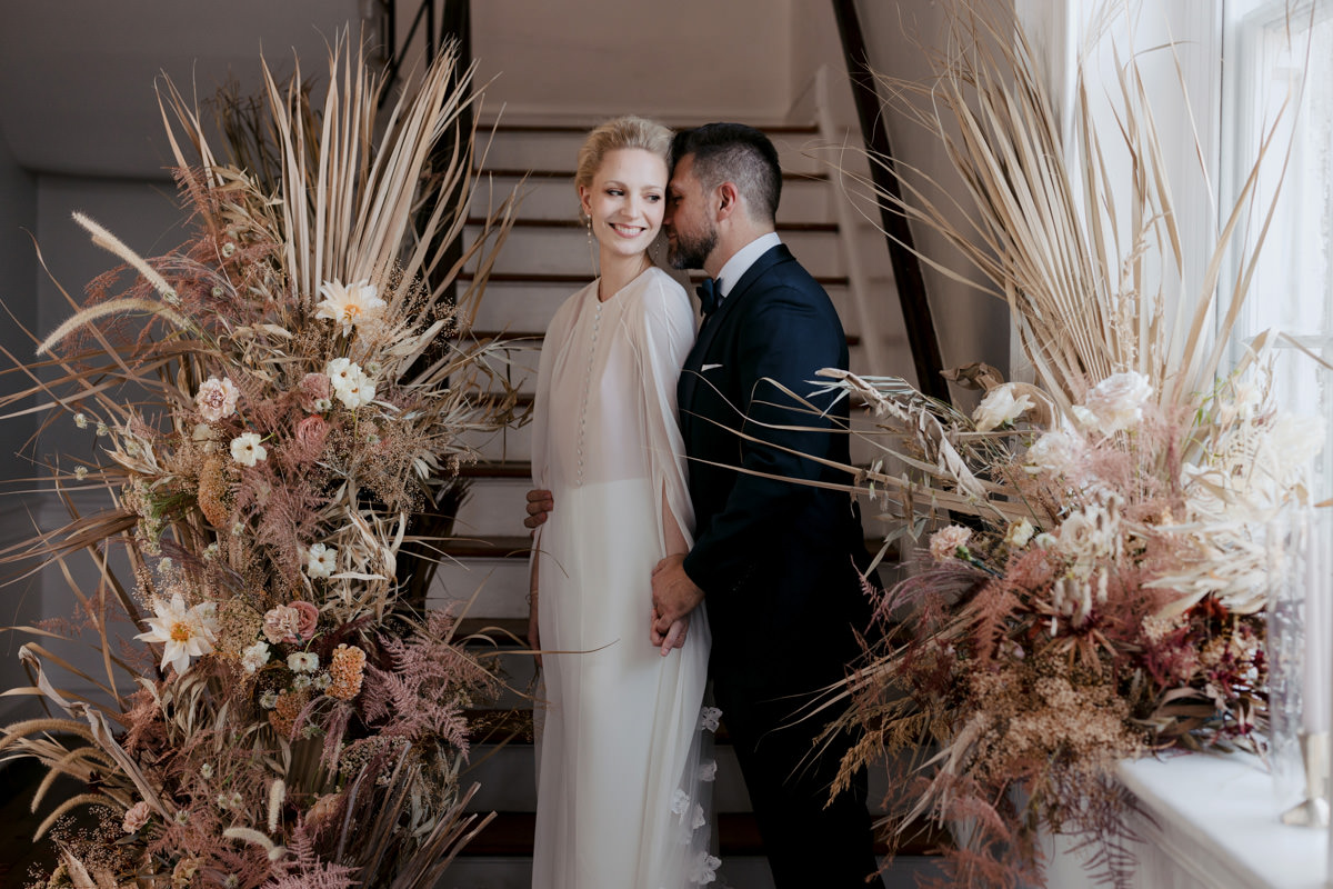 A groom holds his brides hand at the of a staircase surrounded by flowers a wedding planner placed