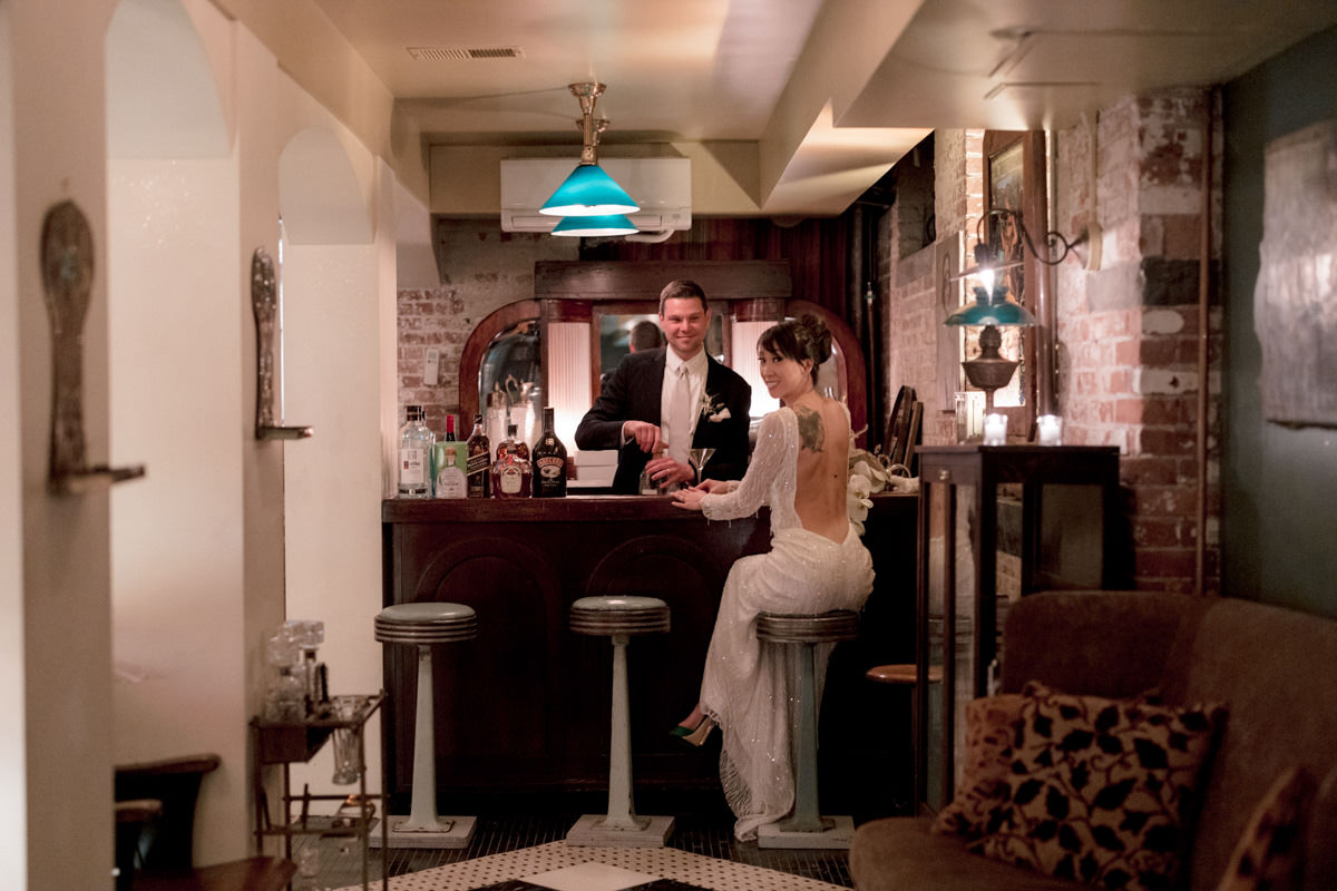 A bride sits at the bar while her groom makes a cocktail