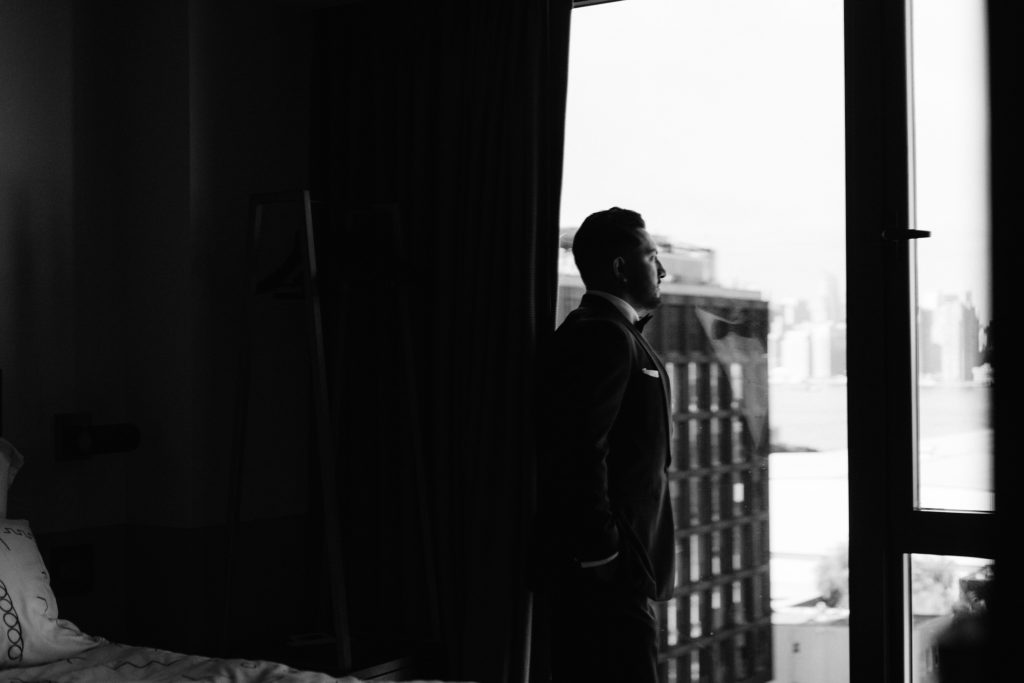 A groom looks out the window of his hotel room.