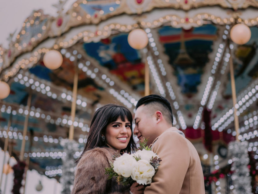 Woman smiles at the camera with man in front of a carousel in Paris during their engagement photos.