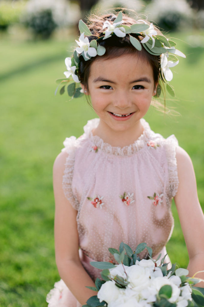 A flower girl poses for the wedding photographer with a big smile.