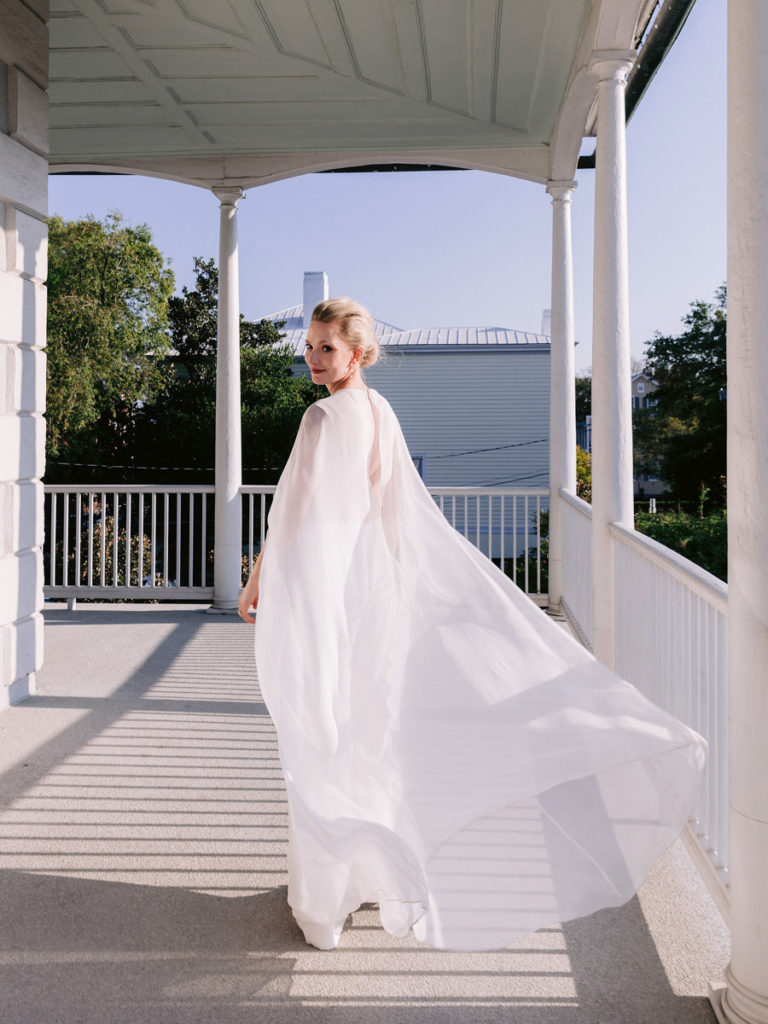 A bride walks on a front porch wearing her wedding dress and cape