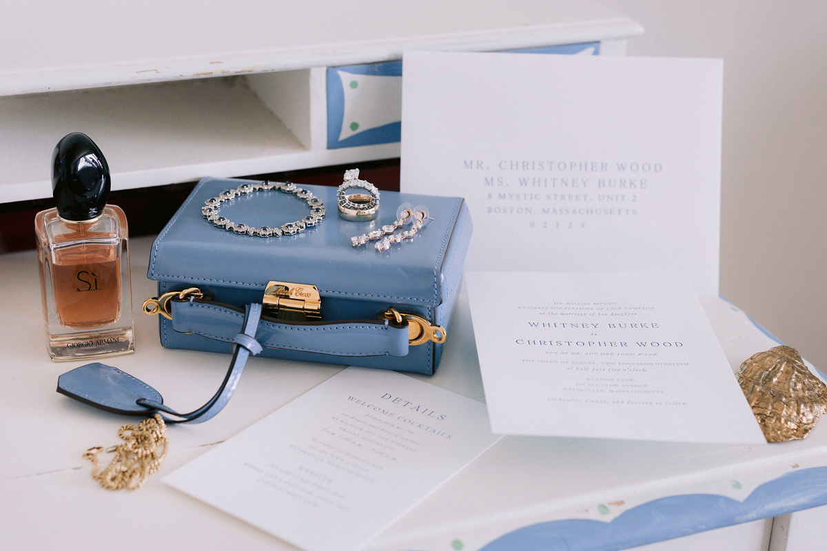 Wedding jewelry and invitations sit on a desk with perfume and a small blue clutch