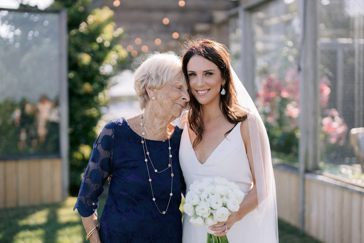 A bride smiles at the camera while her grandmothers head rests on her shoulder.