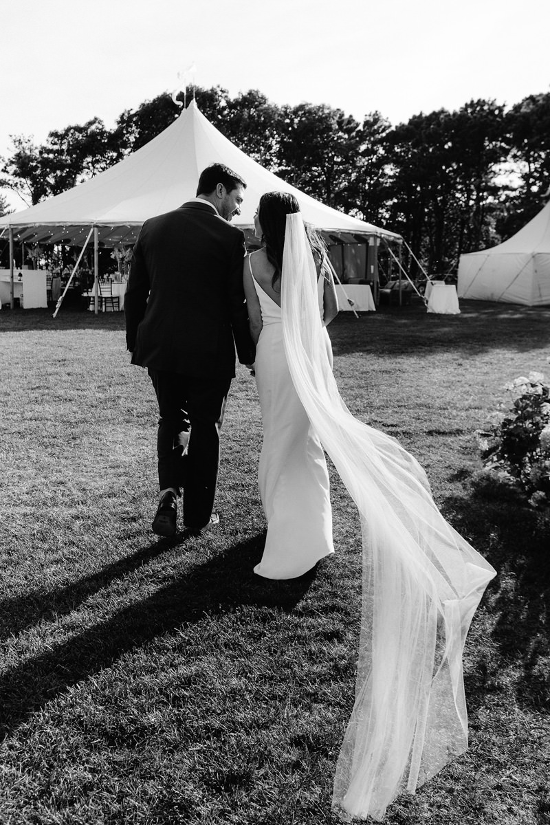 A bride and her groom walking toward their reception with her veil trailing in the wind