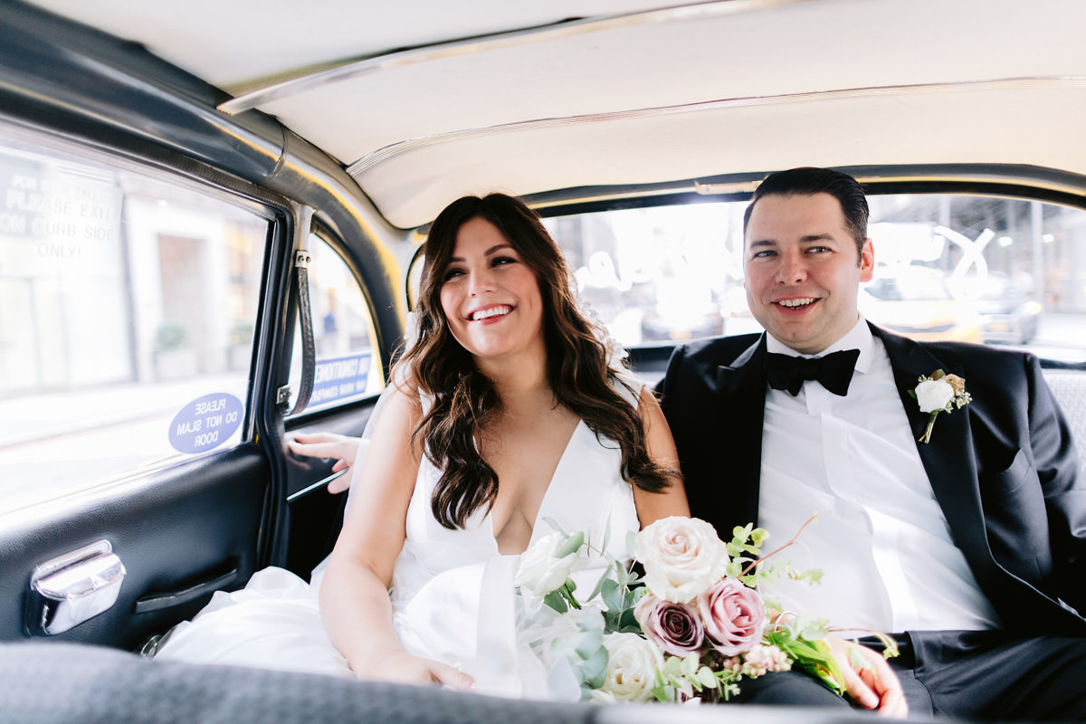 A bride and groom smile in the back of a classic NYC cab with white and pink roses