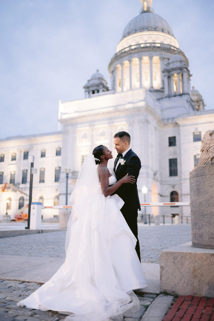 A groom and bride stand outside of a large white building