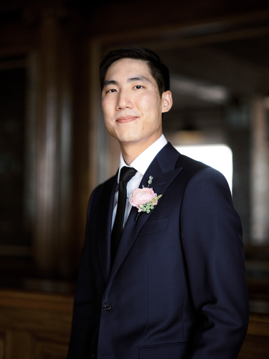 A groom smiles at the camera with a pink ranunculus boutonnière and black tie