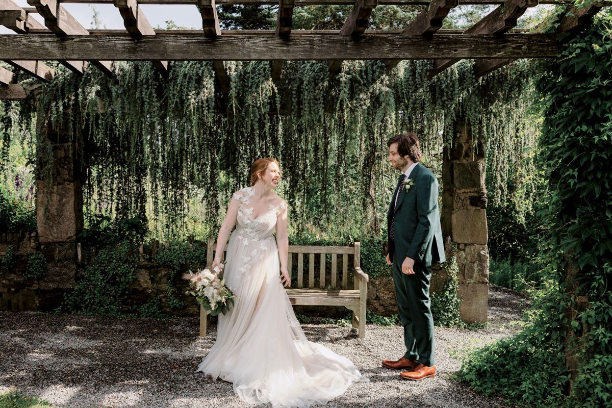 Bride and groom standing, smiling and facing each other, a bit apart; trellis with vines and wooden bench on the background