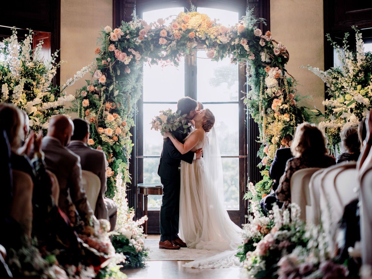Bride and groom kissing under a flowered arch, as people seated watched them.