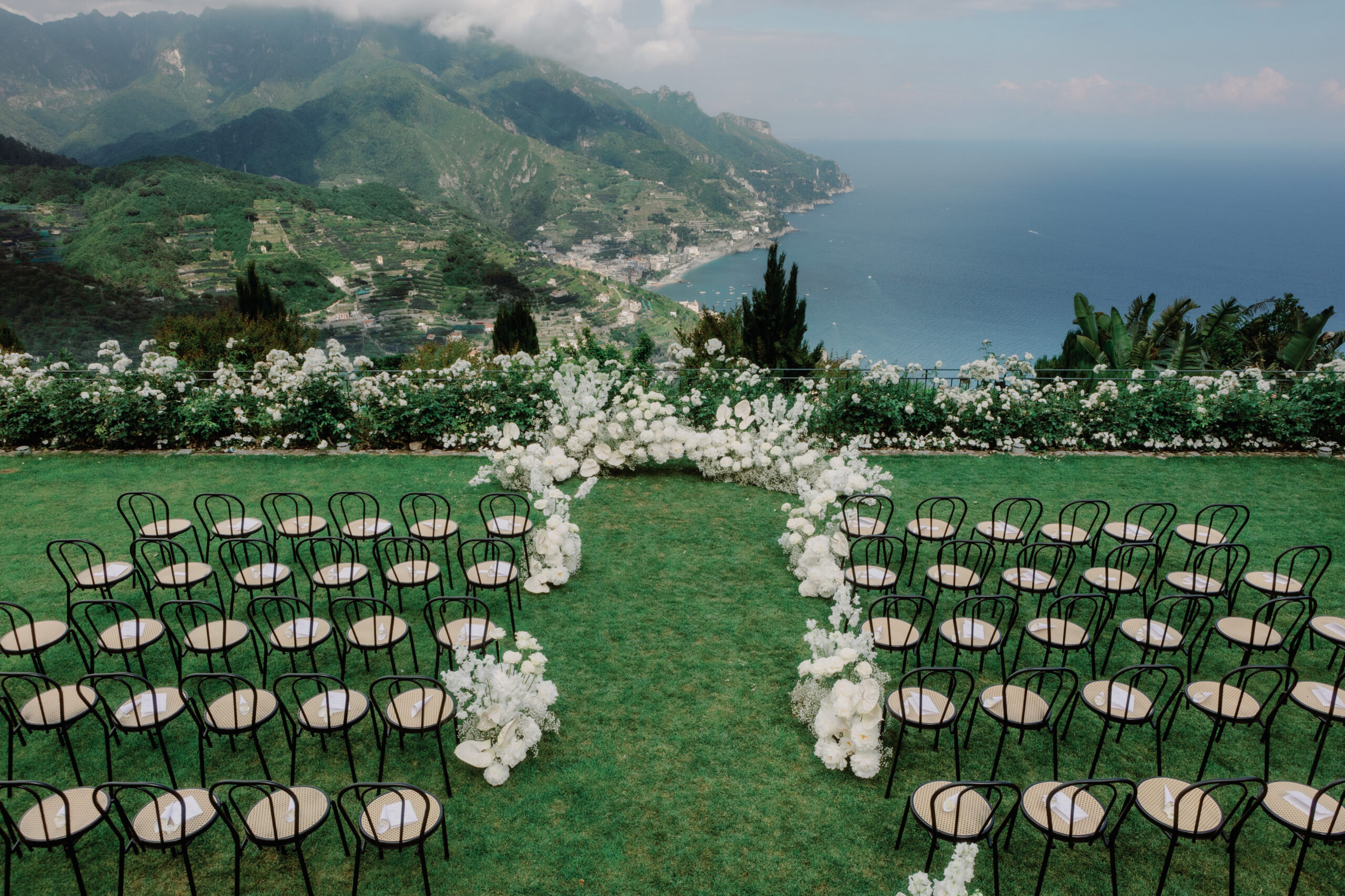 Enchanting wedding ceremony set up at the iconic Caruso, a Belmond Hotel, overlooking the breathtaking Amalfi Coast. The panoramic view features the azure waters, fluffy clouds, and majestic mountains on a beautiful Saturday in June, creating a picture-perfect backdrop for a romantic celebration.