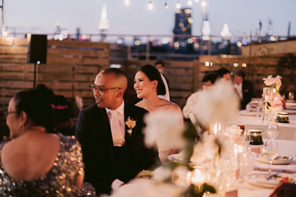 Bride and groom seated with guests on a long table with flower and candle centerpieces; wooden pallet walls on the background