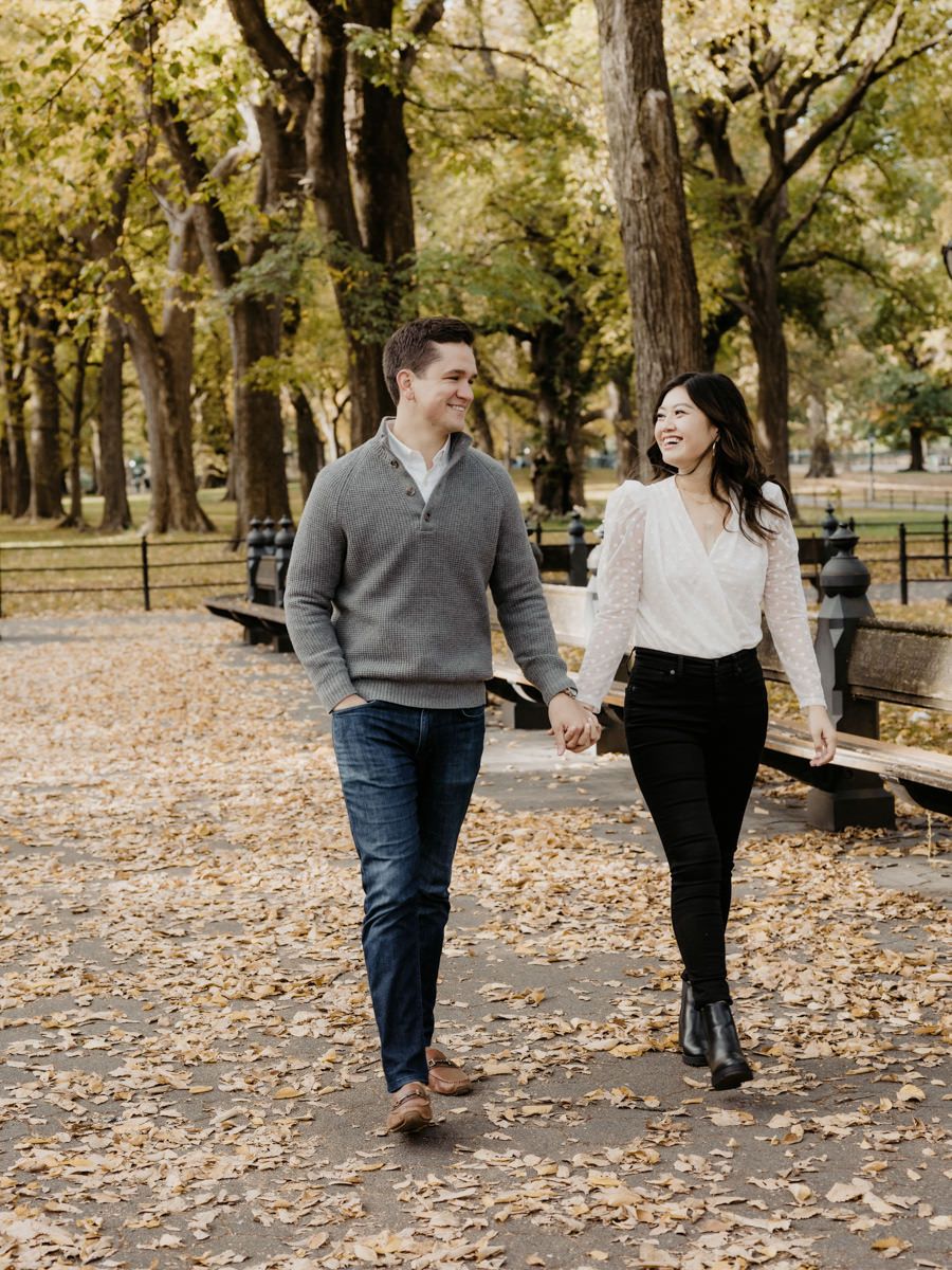 A man and woman holding hands while walking. Trees and fallen leaves on the background