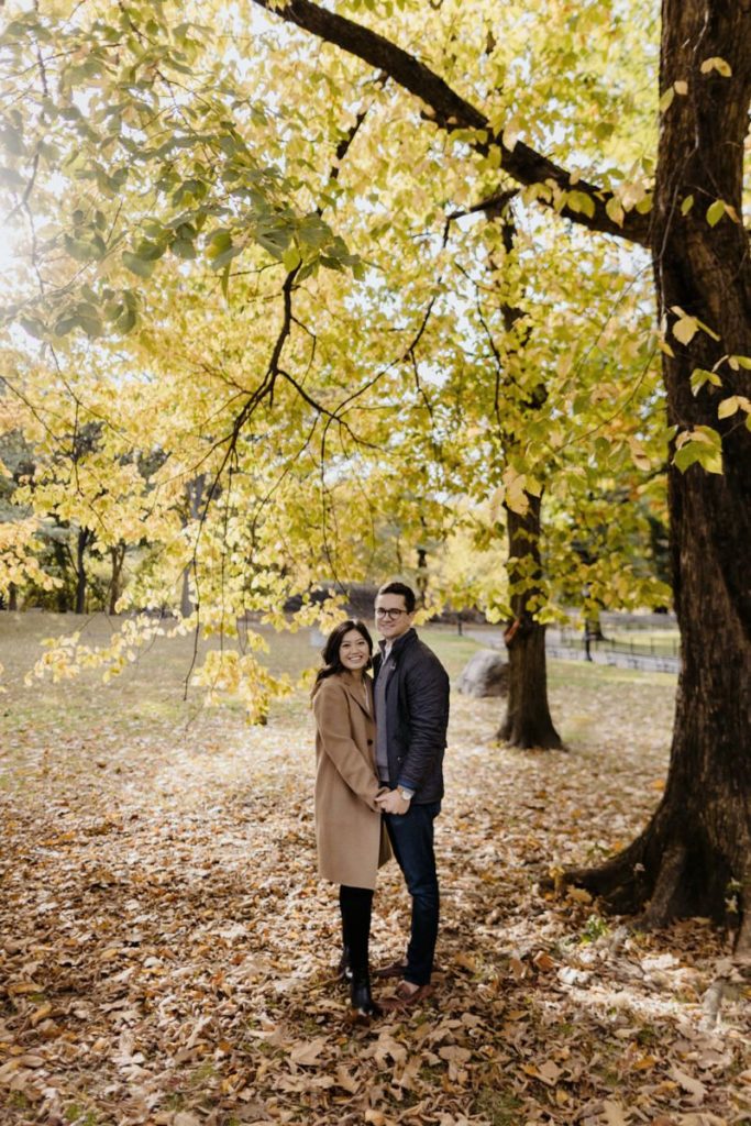 Man and woman standing close to each other, holding hands and smiling. Background is yellow leaf trees & fallen brown leaves