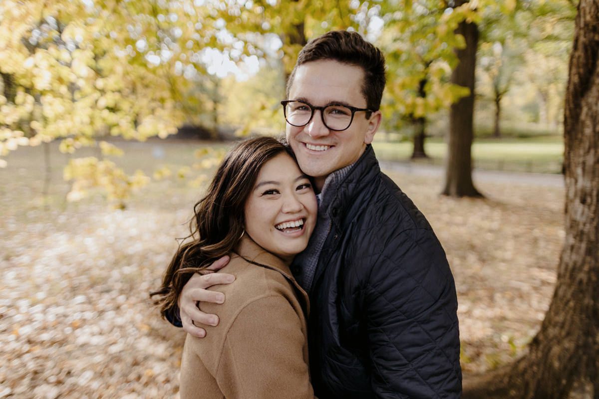 Close-up shot of a man and a woman hugging each other, smiling. Background is yellow leaf trees & fallen brown leaves