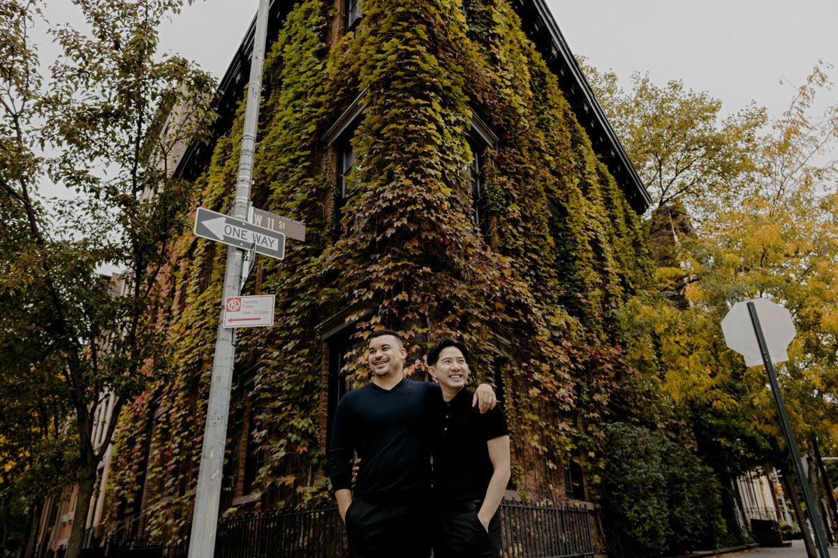 Men couple standing in the middle of a corner street; Background is a building covered with leaves and trees of autumn colors