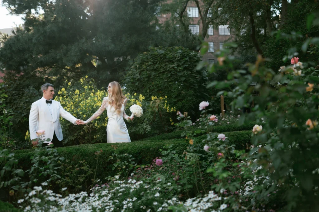 Elegant bride in a satin gown with lace sleeves and a groom in a white tuxedo walking hand-in-hand through a lush garden in Manhattan, surrounded by blooming flowers and timeless greenery. Photographed by Jenny Fu, NYC wedding photographer, specializing in editorial and film photography.