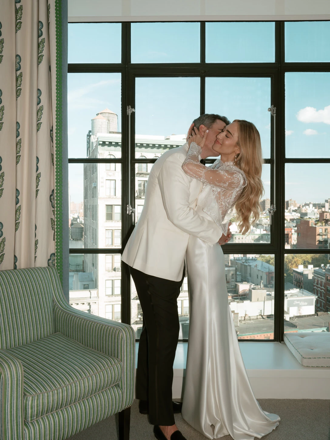 Romantic moment between a bride in a satin gown with lace sleeves and a groom in a white tuxedo, sharing a kiss in front of a floor-to-ceiling window overlooking the iconic Manhattan skyline. Photographed by Jenny Fu, NYC wedding photographer, capturing timeless and elegant celebrations.