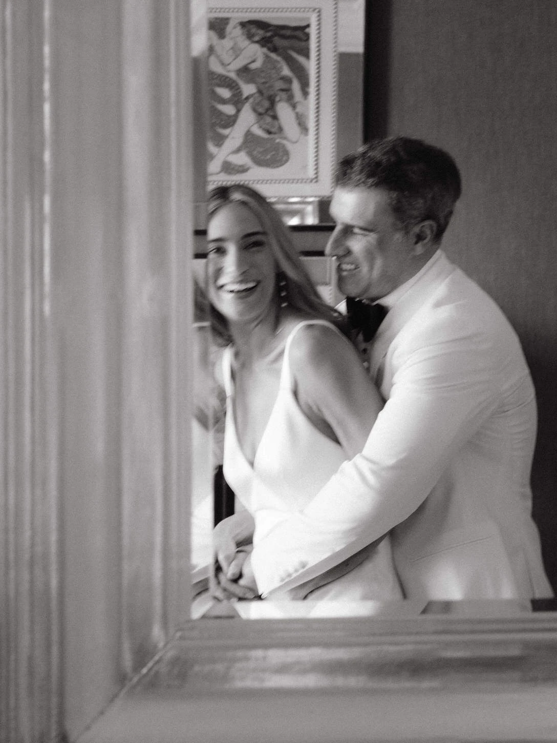 Playful and candid black-and-white moment of a bride in a sleek satin gown laughing as her groom embraces her, framed naturally through a doorway. Photographed by Jenny Fu, NYC wedding photographer, capturing joyful and authentic wedding day memories.