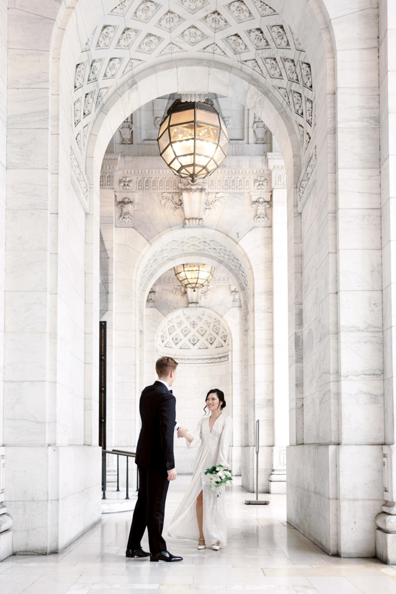 Bride and groom in the middle of New York Public Library's hallway. Large chandeliers at the top