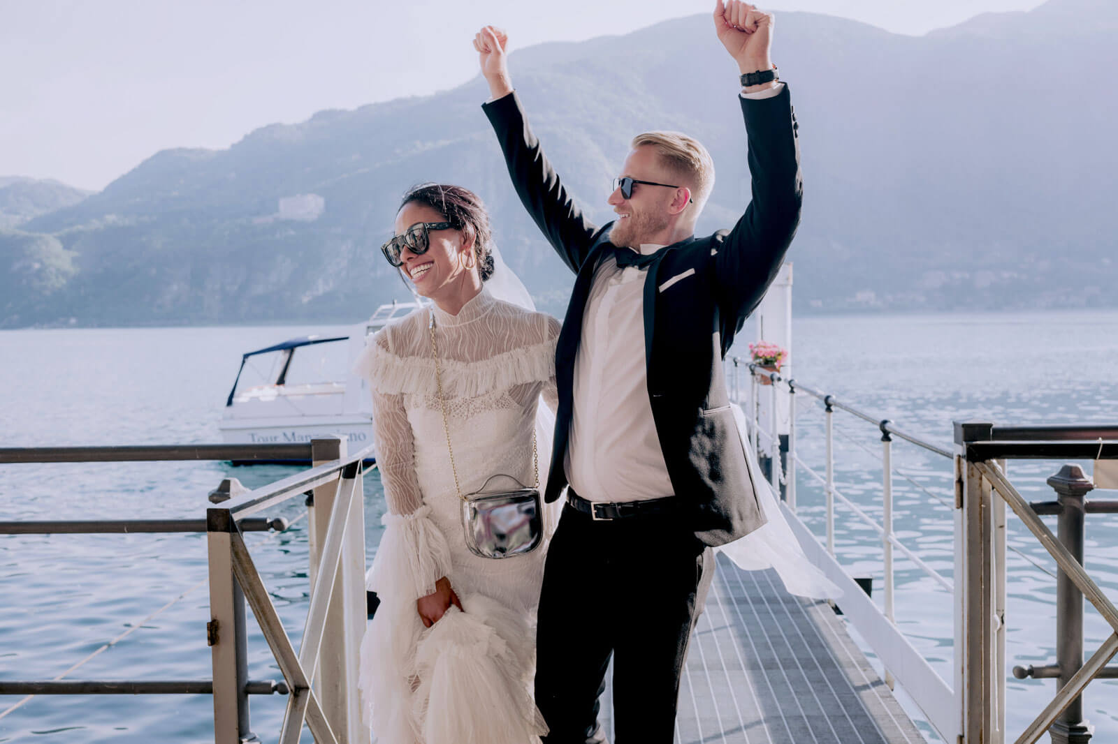 The groom, raising his two arms, is walking with the bride on a dock, with a boat, ocean, and mountain in the background.