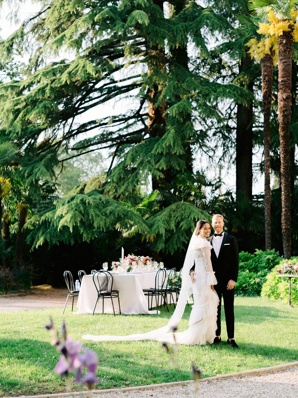 The bride and groom are standing in a garden with a long dining table and large trees in the background.