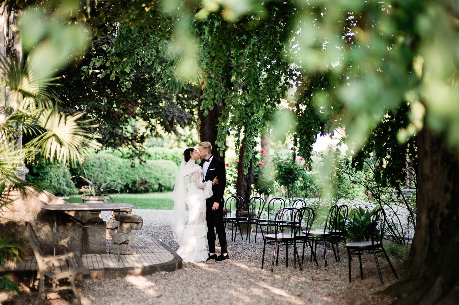 The bride and groom are kissing in a garden full of trees, with chairs on one side and an old table and bench on the other.