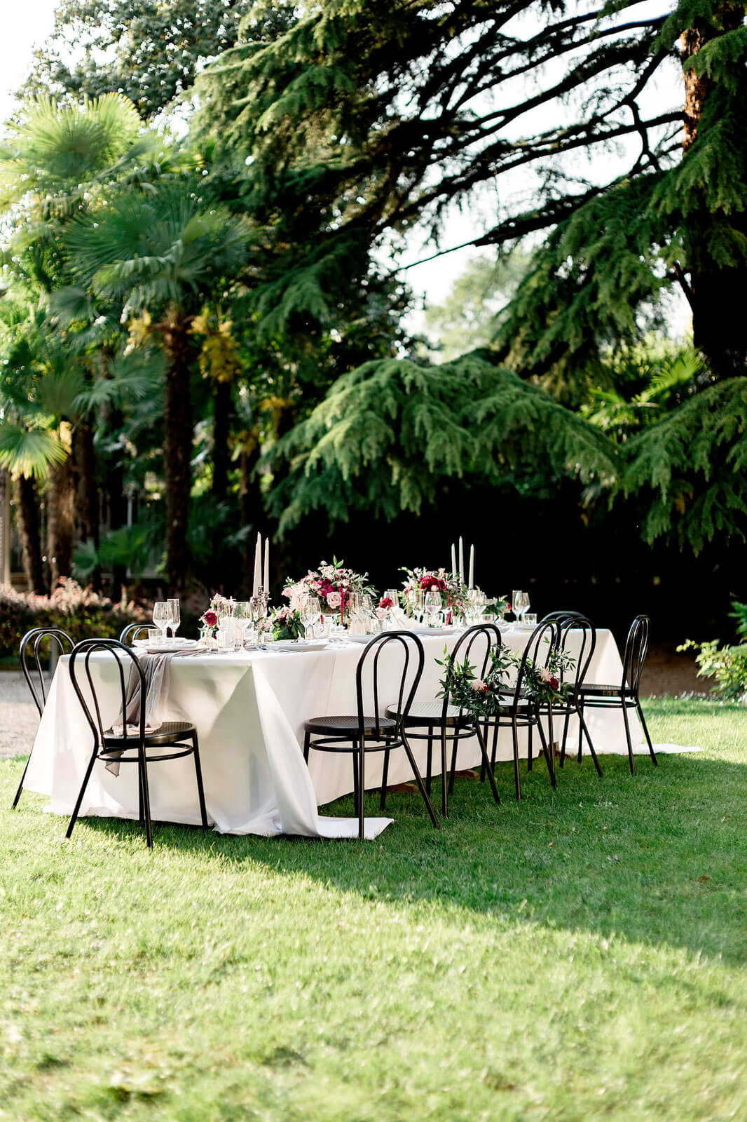 A long dining table and chairs, with white table cloth, flower bouquet and candle centerpieces, with trees on the background