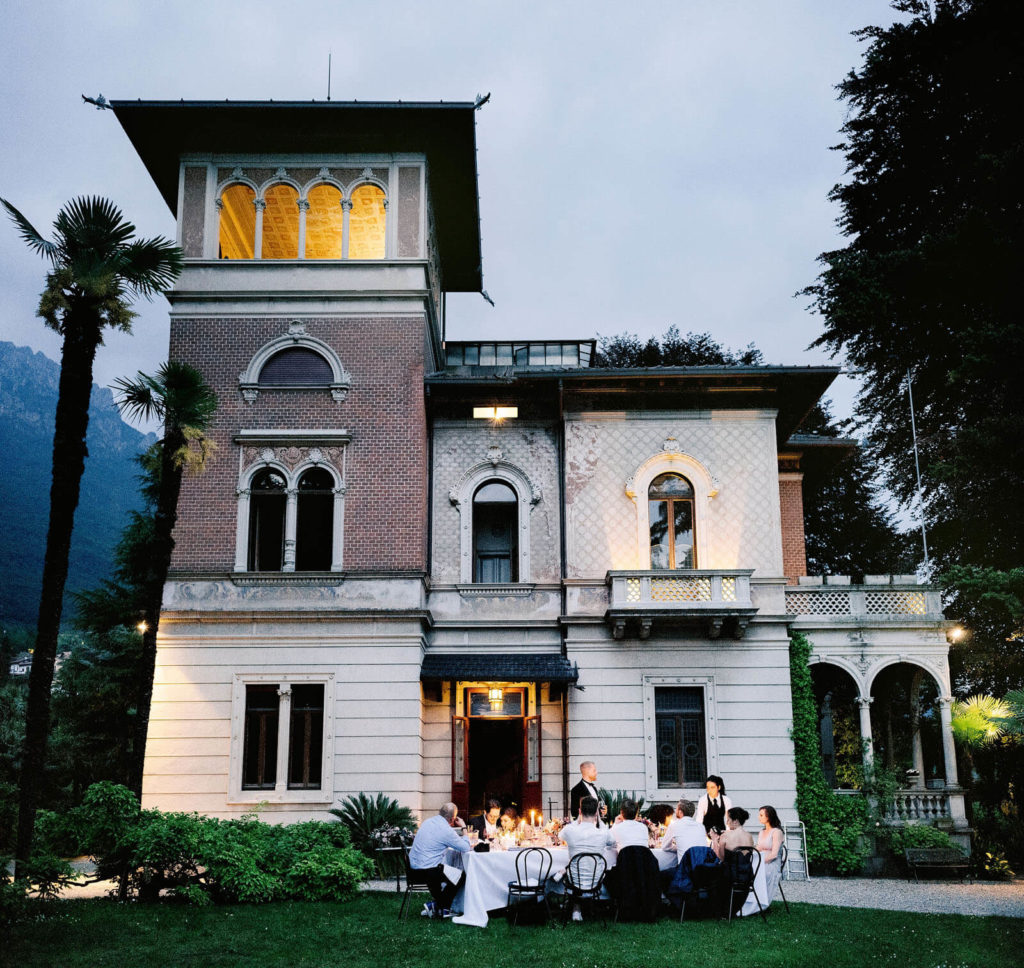 An old three-storey villa with a group of people eating at a long dining table on the front lawn of the villa.