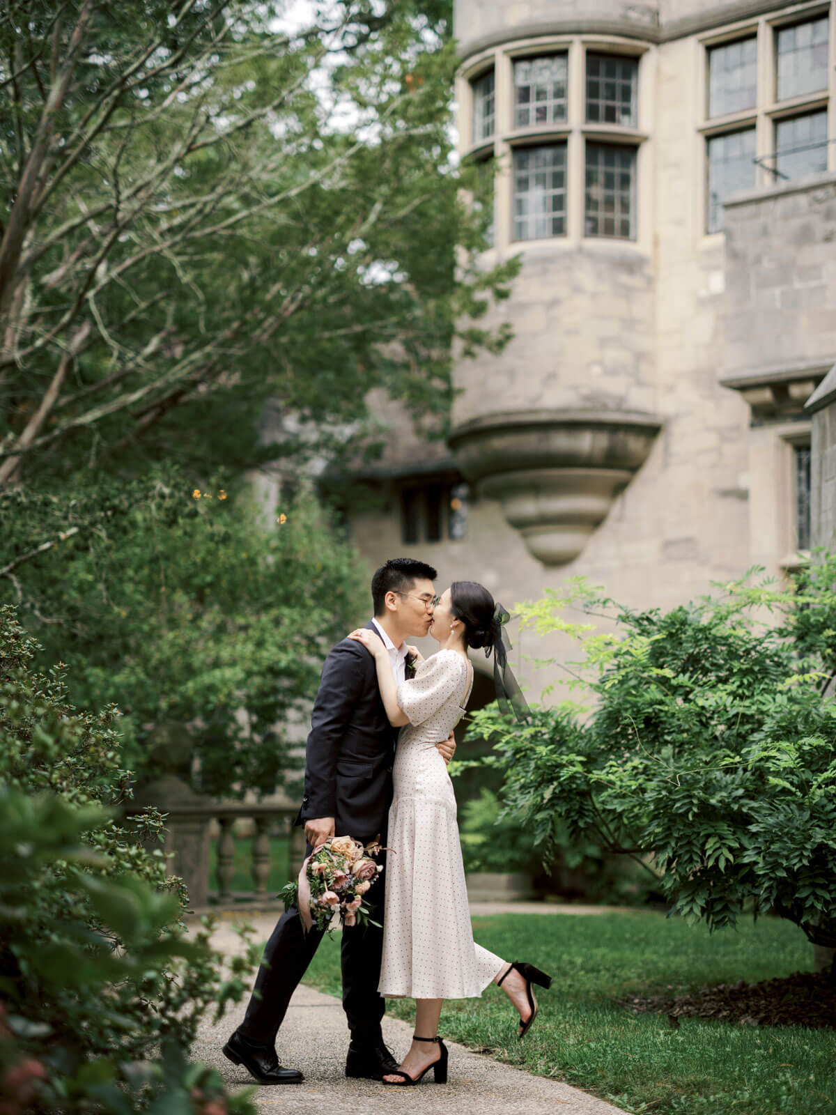 The engaged couple is kissing outside Coe Hall at Planting Fields Arboretum, NY, with lush trees on the side. Image by Jenny Fu Studio