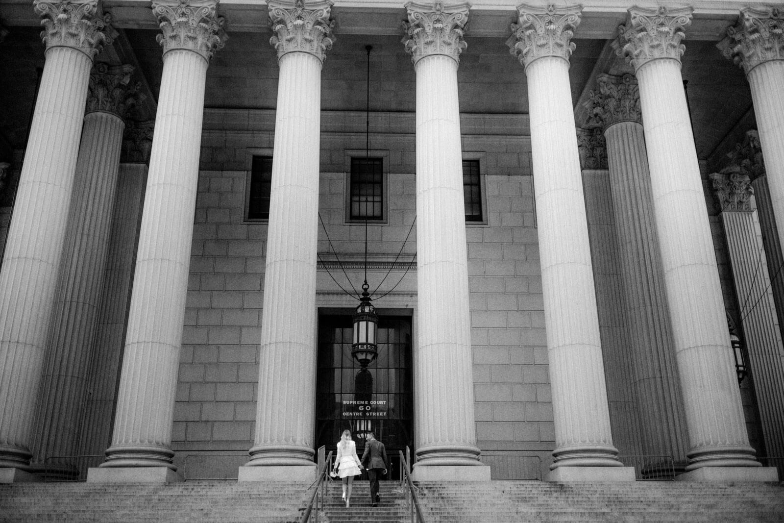 Black and white photo of the bride and the groom climbing up the front staircase of the New York State Supreme Court Building.