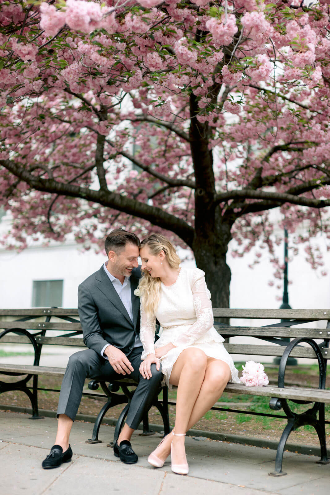 The bride and the groom are sitting on a bench in the park with a cherry blossom tree in the background.