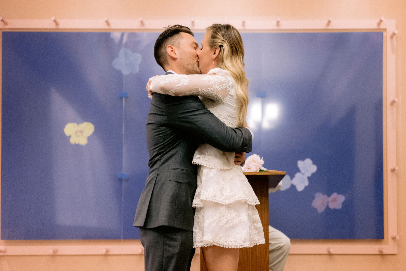 The bride and the groom are kissing in front of the wedding officiant inside the ceremony room at NY City Hall.