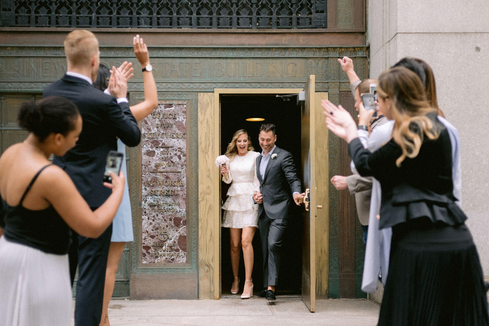 Newlyweds coming out of the New York State Building as a few guests cheer, clap, and take pictures outside.