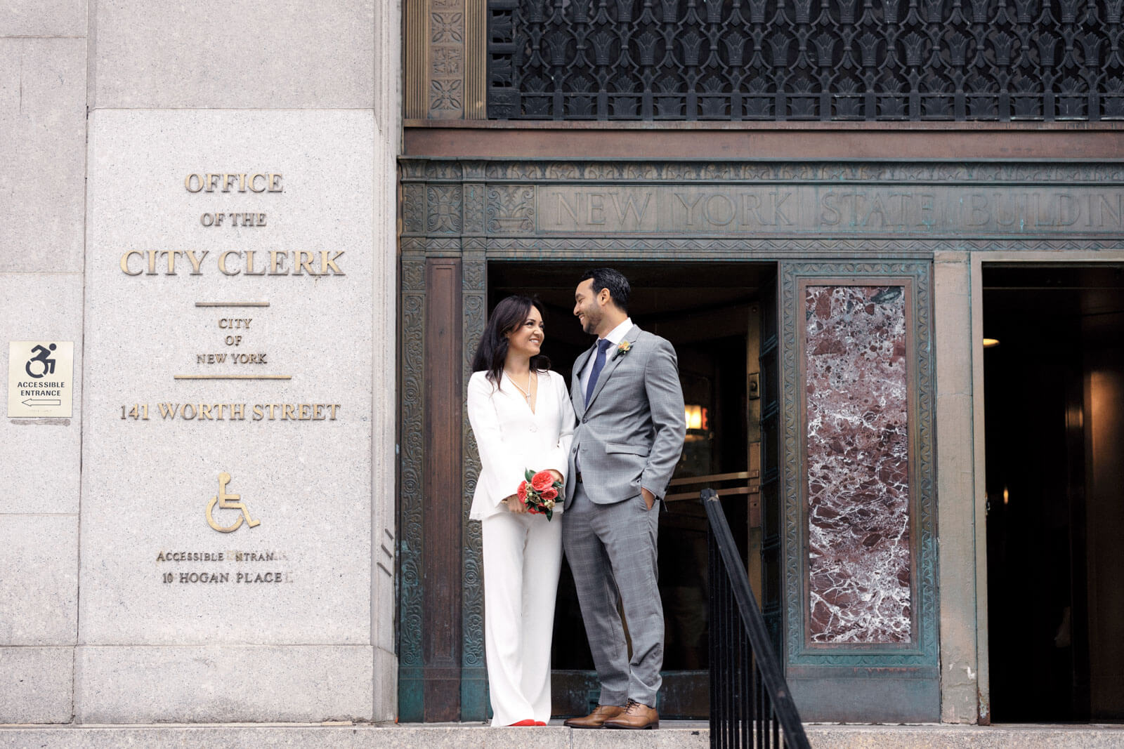 The bride and the groom are standing in front of the Office of the City Clerk Building in 141 Worth Street, New York City.