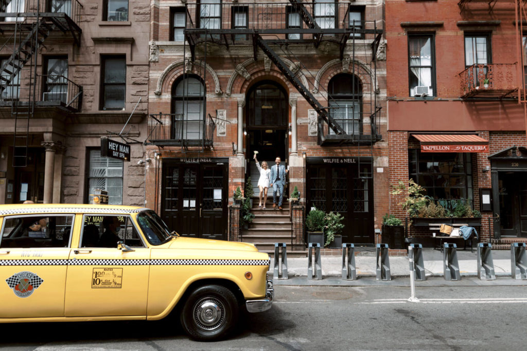 The bride and the groom are standing on the front staircase of Wilfie and Nell Pub NYC. In front is an NYC yellow cab.