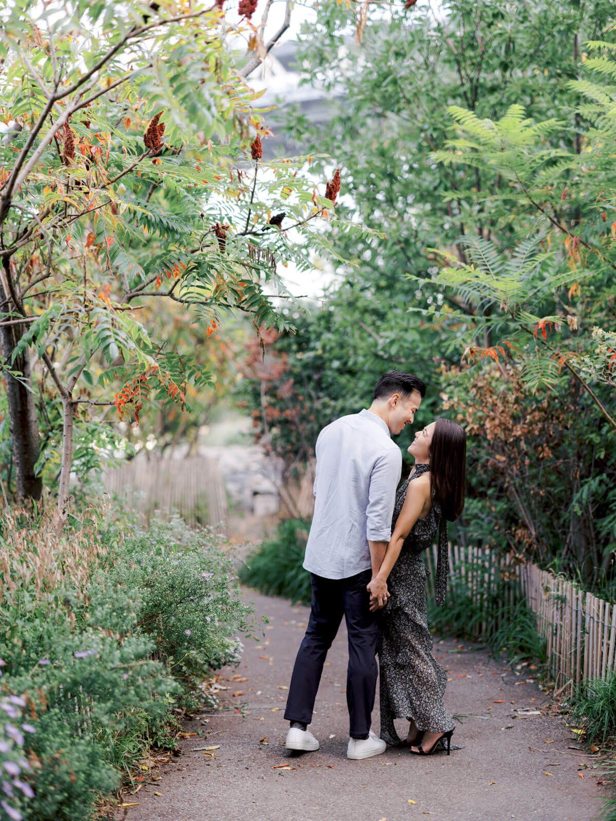 The engaged couple is romantically staring at each other amidst the lush greens at Brooklyn Park, NYC. Image by Jenny Fu Studio