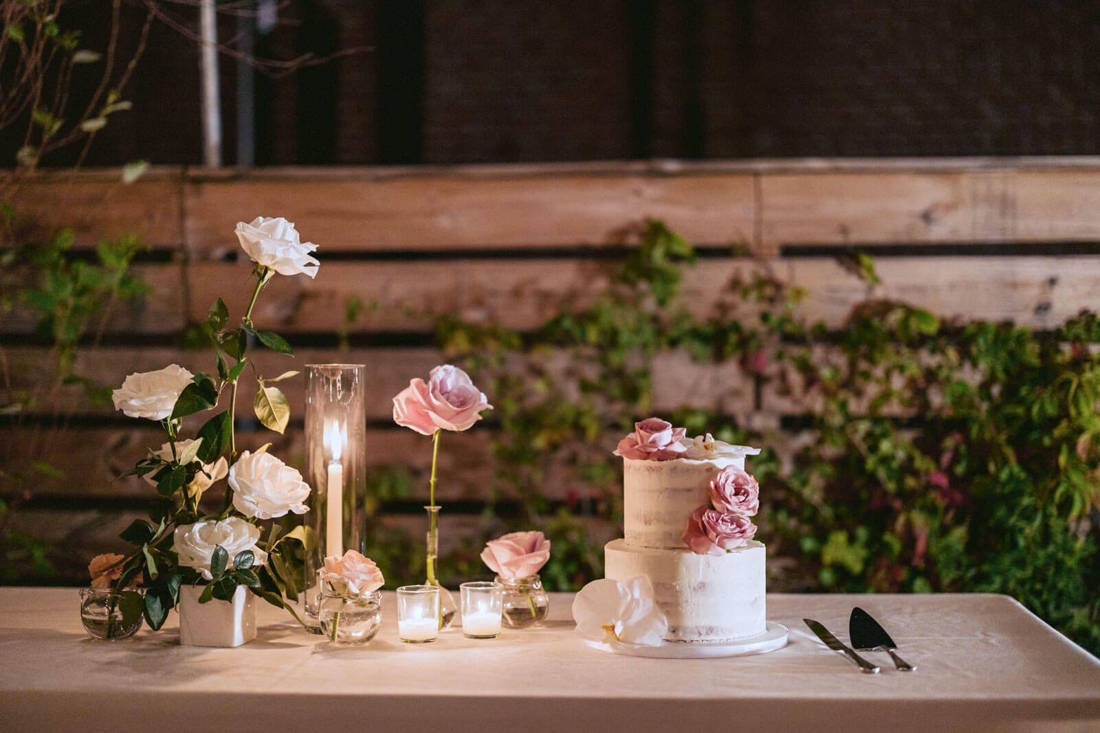 A beautiful two-tiered, naked, small, white wedding cake on a table with candles, pink and white roses and cake cutters in New York City. Image by Jenny Fu Studio