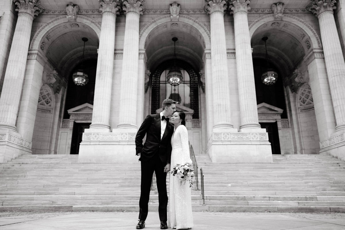 The groom is kissing the bride on the forehead at the grand entrance of the New York Public Library. Image by Jenny Fu Studio